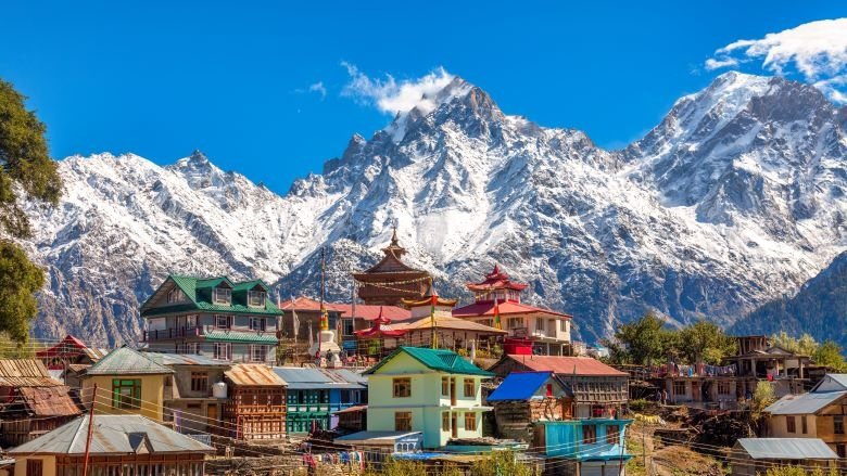 A village with colorful buildings sits on a hill with snow-capped mountain peaks rising behind it